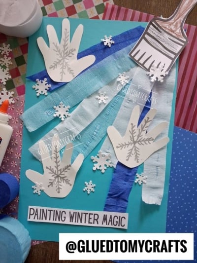 Craft project on blue paper featuring three paper handprints with snowflake designs, streamers, paper snowflakes, a paper paintbrush, and a label reading PAINTING WINTER MAGIC.