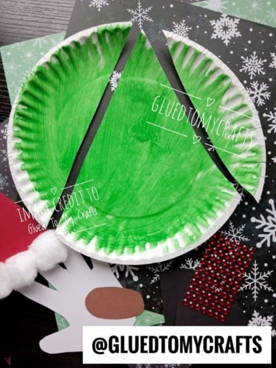A paper plate painted green and cut into three sections to form a Christmas tree, with craft supplies including a handprint cutout, a red hat brim, and red rhinestones on a snowflake background.