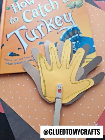 A handmade handprint clothespin turkey rests on a copy of the book How to Catch a Turkey.
