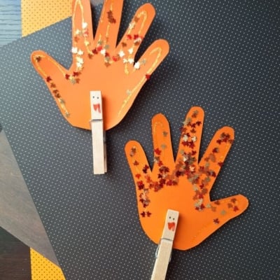 Two orange paper handprint crafts decorated with leaf-shaped glitter and wooden clothespins, set against a dark dotted background.