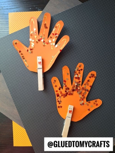 Two orange paper handprint crafts decorated with leaf-shaped glitter and wooden clothespins, set against a dark dotted background.
