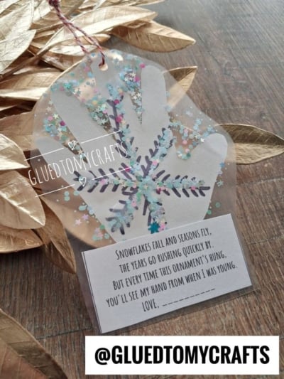 Clear plastic handprint ornament decorated with glitter and a snowflake, featuring a poem about childhood and the brand name GluedToMyCrafts.