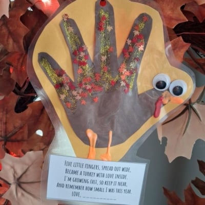 A handprint turkey craft decorated with glitter and autumn leaf cutouts, featuring googly eyes, an orange beak, a red wattle, and a poem about growing up attached at the bottom.
