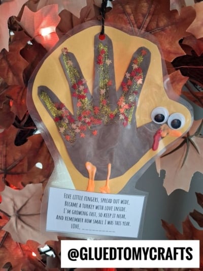 A handprint turkey craft decorated with glitter and autumn leaf cutouts, featuring googly eyes, an orange beak, a red wattle, and a poem about growing up attached at the bottom.