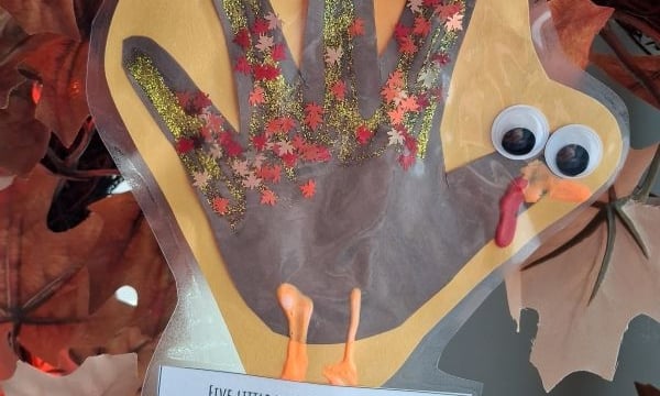A handprint turkey craft decorated with glitter and autumn leaf cutouts, featuring googly eyes, an orange beak, a red wattle, and a poem about growing up attached at the bottom.