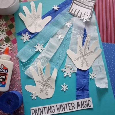 A craft project on light blue cardstock featuring three paper handprints stamped with snowflake designs, streamers, paper snowflakes, and a paper paintbrush cutout, with the text PAINTING WINTER MAGIC at the bottom.