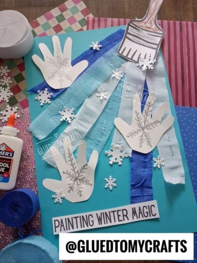 A craft project on light blue cardstock featuring three paper handprints stamped with snowflake designs, streamers, paper snowflakes, and a paper paintbrush cutout, with the text PAINTING WINTER MAGIC at the bottom.