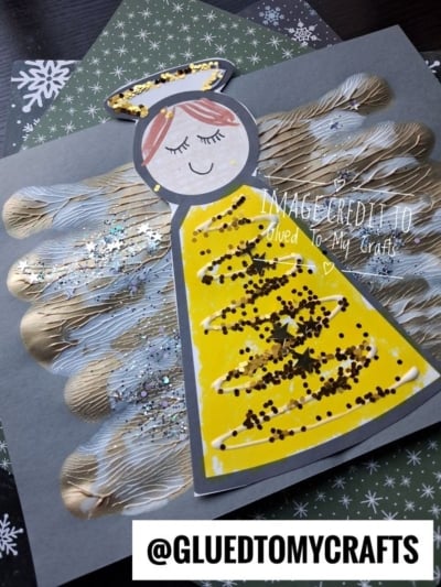 A paper angel craft featuring a handprint-shaped body with gold-painted wings, a yellow triangular dress decorated with glitter, and a paper-cut face with a gold glitter halo.