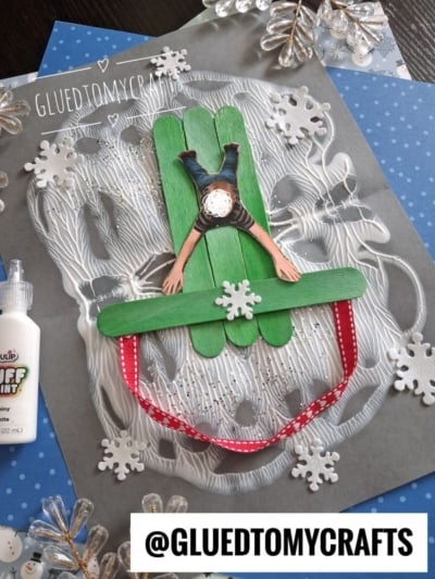 A craft made of green popsicle sticks forming a sled, with a paper cutout of a person lying face down on it, embellished with puffy paint, glitter, and white paper snowflakes.