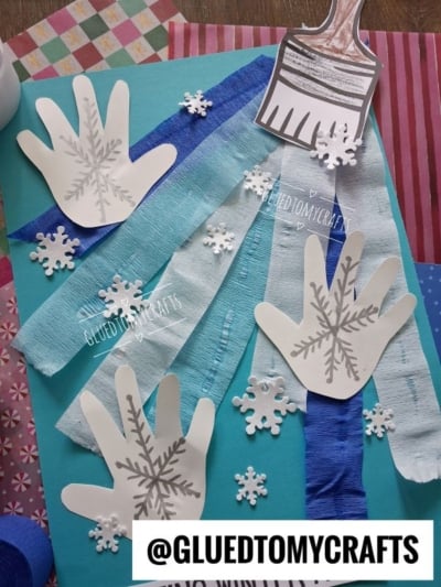 A craft project on blue paper featuring three white handprint cutouts decorated with snowflakes, blue streamers, and a paper paintbrush.