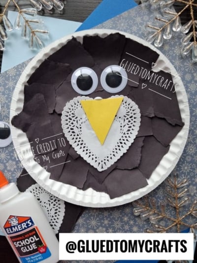 Paper plate craft of an owl made with torn black paper, a white paper doily, a yellow triangle beak, and two googly eyes.