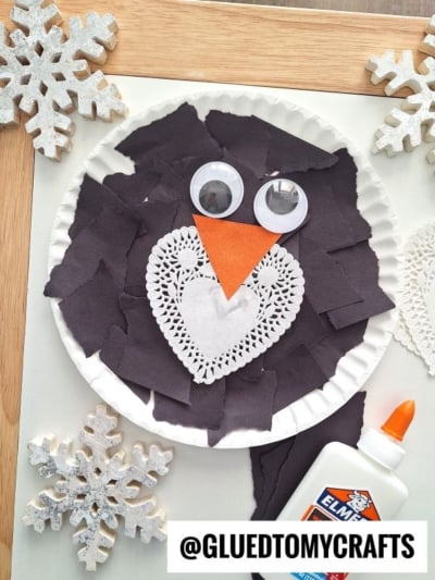 A paper plate craft of a penguin made with torn black paper, googly eyes, an orange paper beak, and a heart-shaped doily on its chest.