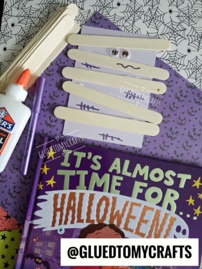 A popsicle stick mummy on purple spiderweb paper, surrounded by a book titled Its Almost Time for Halloween, craft glue, and a purple marker as part of a Halloween craft for kids.