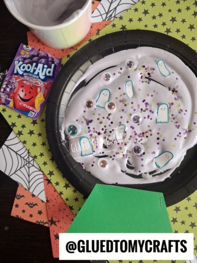 A black plate filled with white slime decorated with googly eyes, ghost cutouts, and colorful glitter, sitting next to a packet of grape Kool-Aid and patterned Halloween paper for fun Halloween crafts for kids.