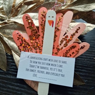 A turkey craft made from a painted popsicle stick and paper handprints covered in glitter, with an attached card that reads, A thanksgiving craft with love to share, to show you just how much I care.