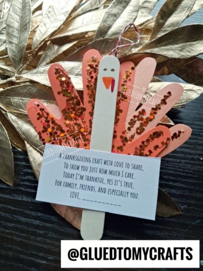 A turkey craft made from a painted popsicle stick and paper handprints covered in glitter, with an attached card that reads, A thanksgiving craft with love to share, to show you just how much I care.