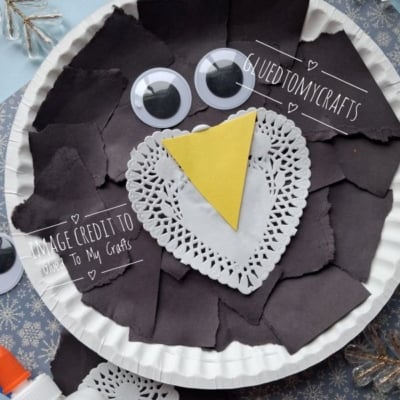 A paper plate craft of a penguin made with black paper scraps, a white paper doily, a yellow triangle beak, and two googly eyes.
