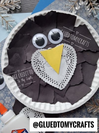 A paper plate craft of a penguin made with black paper scraps, a white paper doily, a yellow triangle beak, and two googly eyes.